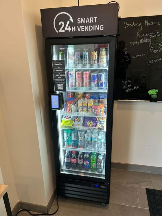 A second managed micro market installed in a different location, alongside a chalkboard where customers have written 'Vending Machine Requests' with handwritten asks, proof that demand at the spot pre-dates the install.