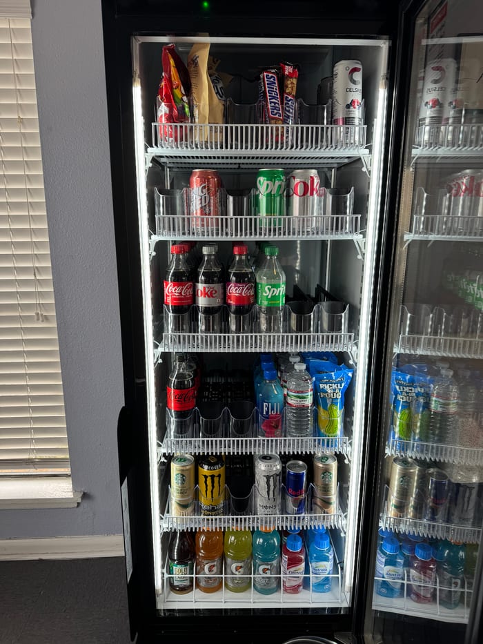 Interior close-up of the machine, six shelves visible, top to bottom: snacks and candy, Coca-Cola cans, Coke and Sprite bottles, Fiji and Pickle Pouch, Monster and Starbucks energy, Gatorade.