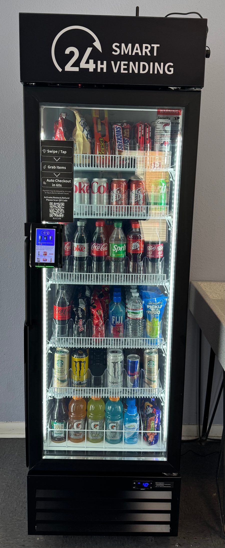 A floor-to-ceiling managed micro market unit installed in a workplace, fully stocked with sodas, energy drinks, water, snacks, and grab-and-go essentials. Branded '24H Smart Vending' with a built-in tap-to-pay screen on the side.